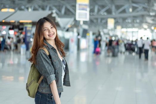 Happy beauty Asian woman traveling and holding suitcase in airport terminal with crowd people passengers background. People and Lifestyle concept. Journey around the world theme. Cheerful tourist girl