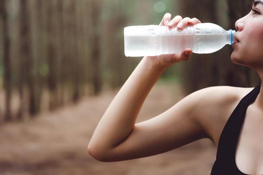 Close up of Beauty Asian woman  drinking purify water by right hand after exercise in forest. Thirsty girl in black sport wear. Workout and Sports concept. People and Outdoor Nature theme.