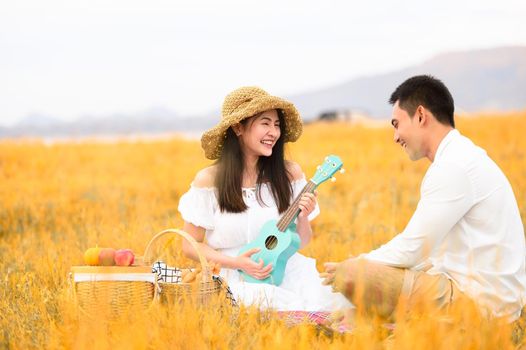 Two Asian young couples in autumn meadow field doing picnic in honeymoon trip in white clothes, ukulele guitar and fruits basket. People lifestyle and wedding concept. Nature and travel day concept.