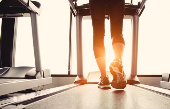 Lower body at legs part of Fitness girl running on running machine or treadmill in fitness gym with sun ray. Warm tone. Healthy and Exercise activity concept. Workout and  Strength training theme.