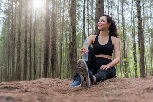 Beauty Asian sport woman resting and holding drinking water bottle and relaxing in middle of forest after tired from jogging. Girl sitting and looking attraction view. Workout concept. Lifestyle theme