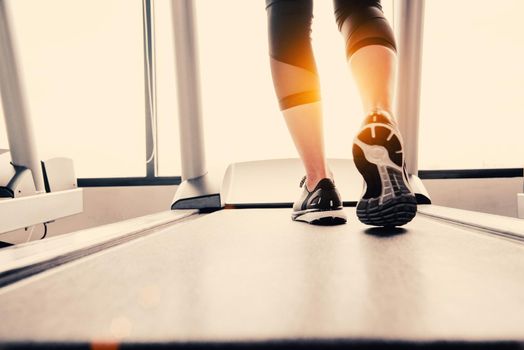 Lower body at legs part of Fitness girl running on running machine or treadmill in fitness gym with sun ray. Warm tone. Healthy and Exercise activity concept. Workout and  Strength training theme.