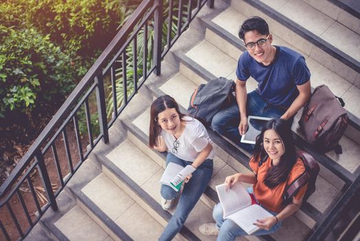 Three Asian young campus students enjoy tutoring and reading books together at stair. Friendship and Education concept. Campus school and university theme. Happiness and funny of learning in college.