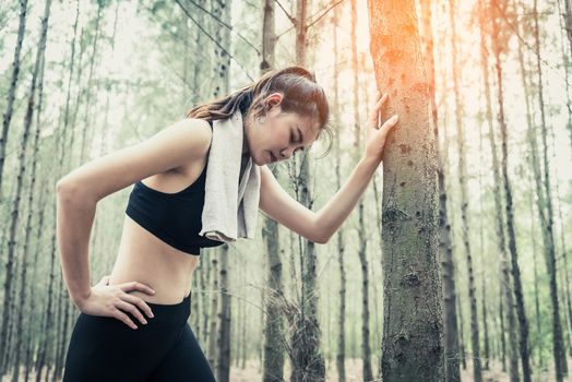 Asian beauty woman tiring from jogging in forest. Lean on tree. Towel and sweat elements. Sport and Healthy concept. Jogging and Running concept. Relax and waist pain theme. Outdoors activity theme.