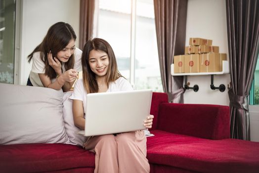 Two young freelancer Asian girl businesswomen private working at home office with laptop computer sitting on sofa. Packaging delivery online shopping service and marketing advertising to customer