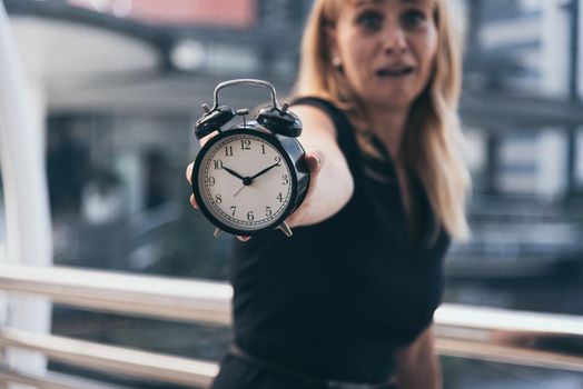 Business women show alarm clock and shocked with late in rush hours when going to work in city urban background. Deadline and wake up late. People lifestyle and daily life planner concept. 