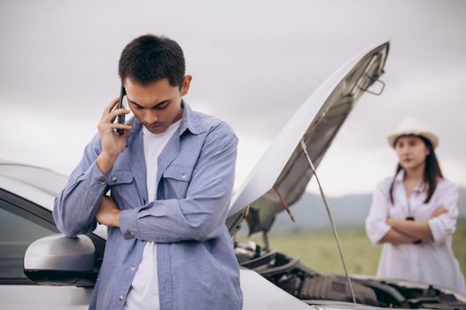 Asian man calling car maintenance service with his girlfriend looking at him background between long road trip. Car broken in mountain meadow with wife. People and transportation concept. Car repair