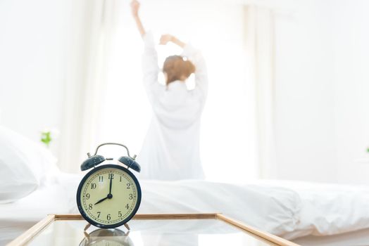 Back view of woman stretching in morning after waking up on bed near window with alarm clock. Holiday and Relax concept. Lazy day and Working day concept. Office woman and worker in daily life theme