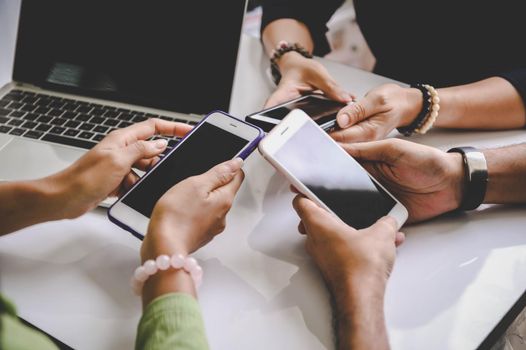 Group of young hipsters holding mobile phones in hands at office with laptop computer background. Friends having fun together with smartphone. Social distancing and work from home concept.