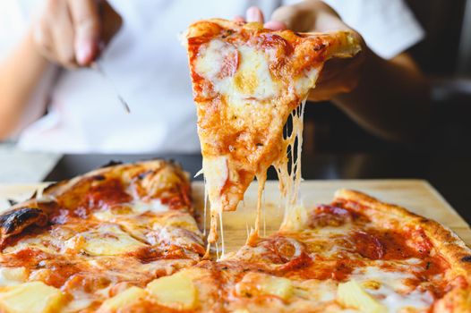 Wood fire cheesy pizza slice in hand. Traditional Hawaiian and Salami pizza savory dish flavour of origin Italian restaurant. Wood-Fired Oven Pizza with people holding and cutting knife background