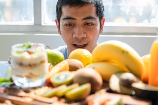 Asian man surprising when seeing many fruit yogurt with granola on table in kitchen background. Food and beverage ingredient concept. Happy vegetarian man hungry and ready to eat vegetable vitamin c