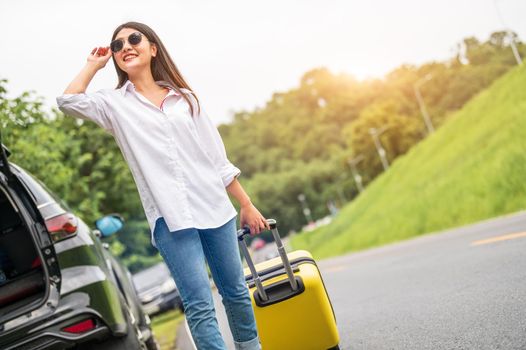 Beauty Asian woman with sunglasses dragging yellow suitcase baggage alone on road trip. People lifestyle and vacation concept. Nature and summer background. Girl having car adventure transportation