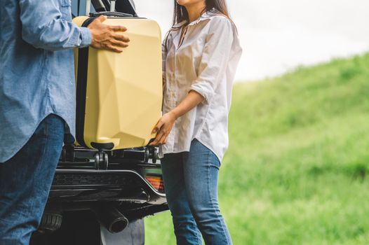 Asian man helping woman to lifting suitcase from car during travel in long weekend. Couple have road trip in vacation with yellow luggage. People lifestyle and transportation concept. Nice guy theme