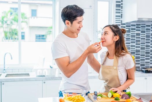 Asian boyfriend feeding fruit and vegetable to girlfriend to each other during preparing for dinner in home kitchen. Couple and Family concept. Honeymoon and Holidays. Man and woman cooking happily