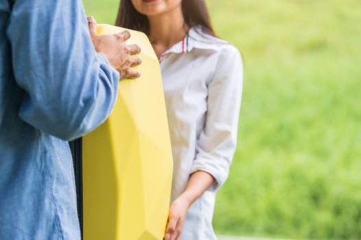 Asian man helping woman to lifting suitcase from car during travel in long weekend. Couple have road trip in vacation with yellow luggage. People lifestyle and transportation concept. Nice guy theme