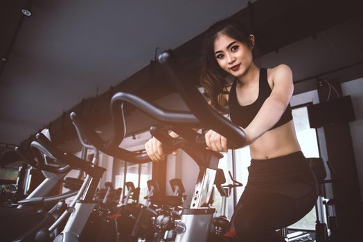 Asian fitness woman on bicycle doing spinning at gym. Beautiful young female work out on gym bike. Medical and Healthy concept. Workout and Cardio theme.