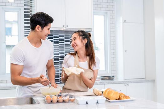 Happy Asian couples cooking and baking cake together in kitchen room. Man and woman looking to tablet follow recipe step at home. Love and happiness concept. Sweet honeymoon and Valentine day theme