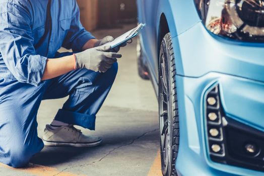 Asian car mechanic technician holding clipboard and checking to maintenance vehicle by customer claim order in auto repair shop garage. Wheel tire repair service. People occupation and business job
