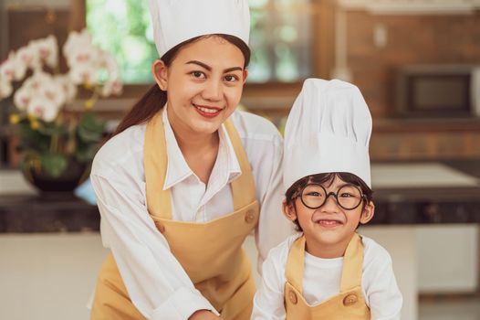 Portrait cute little Asian boy chef with eyeglasses and his mother looking to camera in home cooking kitchen happily and funny. People lifestyles and Family. Homemade food ingredient making concept