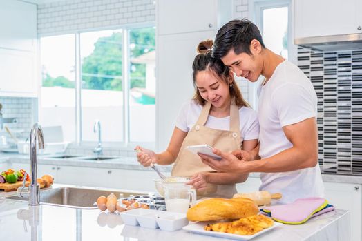 Happy Asian couples cooking and baking cake together in kitchen room. Man and woman looking to tablet follow recipe step at home. Love and happiness concept. Sweet honeymoon and Valentine day theme