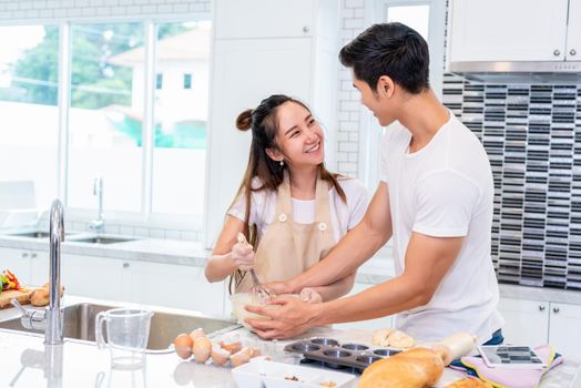 Happy Asian couples cooking and baking cake together in kitchen room. Man and woman looking to tablet follow recipe step at home. Love and happiness concept. Sweet honeymoon and Valentine day theme