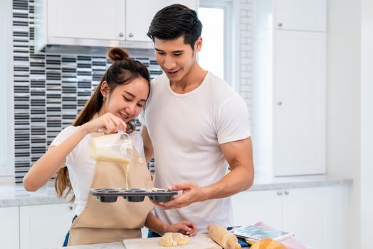 Happy Asian couples cooking and baking cake together in kitchen room. Man and woman looking to tablet follow recipe step at home. Love and happiness concept. Sweet honeymoon and Valentine day theme