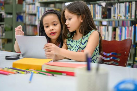 Two little happy cute girls playing on a tablet PC computing device in library at school. Education and self learning wireless technology concept. People lifestyles and friendship. Preschool children