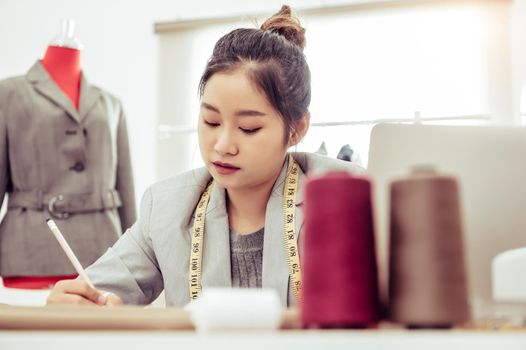 Attractive Asian female fashion designer working in home office workshop. Stylish fashionista woman creating new cloth design collection. Tailor and sewing. People lifestyle and occupation concept