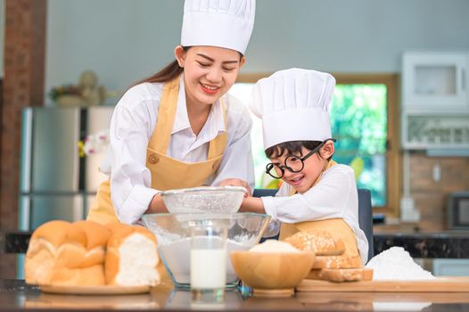 Cute little Asian boy and beautiful mother sifting dough flour with sifter sieve colander in home kitchen on table for prepare to baking bakery and cake. Thai kids playing with flour as chef funny