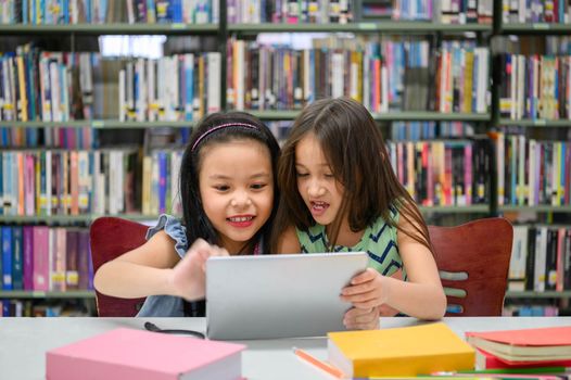 Two little happy cute girls playing on a tablet PC computing device in library at school. Education and self learning wireless technology concept. People lifestyles and friendship. Preschool children