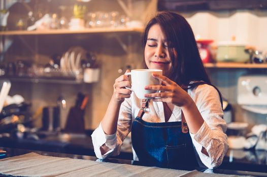 Beauty Asian female barista holding cup of coffee and tasting brewed coffee  with cafe restaurant background. Waitress smelling coffee at cafeteria. Food and drink concept. People lifestyle