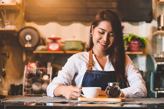 Asian female barista making cup of coffee. Young woman holding white coffee cup while standing behind cafe counter bar in restaurant background. People lifestyles and Business occupation concept.