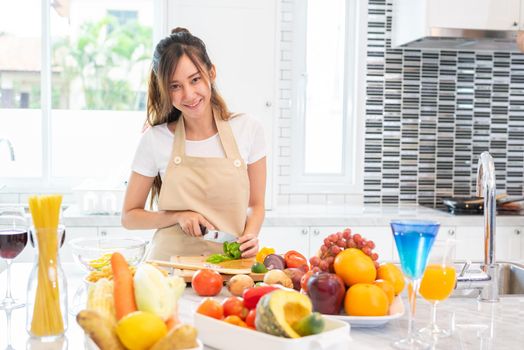 Asian beauty woman cooking and slicing vegetable in kitchen room with full of food and fruit on table for party. Holiday and Happiness concept. People and lifestyles concept. Girl looking at camera