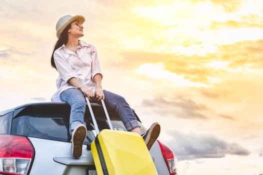 Happy Asian woman on top of car with luggage bag. Girl sitting on roof and looking sunset before night in evening. People lifestyle in long vacation trip concept. Nature and transportation vehicle.