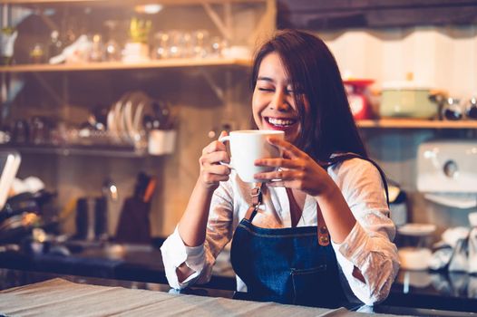 Happy Asian female barista holding cup of coffee and tasting brewed coffee from herself with cafe restaurant background. Waitress at cafeteria. Food and drink. People lifestyle and occupation concept.