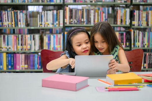 Two little happy cute girls playing on a tablet PC computing device in library at school. Education and self learning wireless technology concept. People lifestyles and friendship. Preschool children