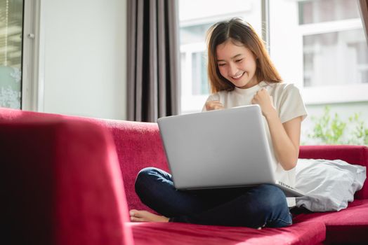 Young woman surfing the internet by laptop on red sofa and glad in successful trading in her house. Selling and online shopping concept. Merchant and marketing concept. Happiness of new trader theme