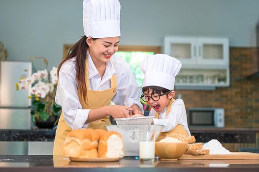 Cute little Asian boy and beautiful mother sifting dough flour with sifter sieve colander in home kitchen on table for prepare to baking bakery and cake. Thai kids playing with flour as chef funny