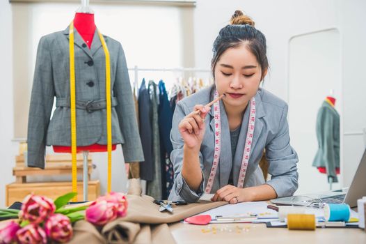 Attractive Asian female fashion designer working in home office workshop. Stylish fashionista woman creating new cloth design collection. Tailor and sewing. People lifestyle and occupation concept