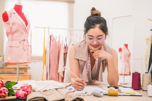 Attractive Asian female fashion designer working in home office workshop. Stylish fashionista woman creating new cloth design collection. Tailor and sewing. People lifestyle and occupation concept
