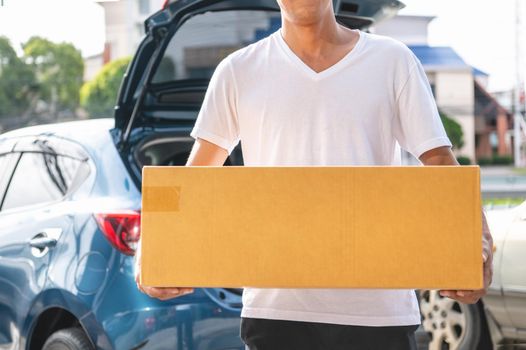 Closeup of happy Asian man delivering cardboard box to customers via private car. People lifestyles and business occupation concept. Young male courier in casual clothes. Shipping and packaging