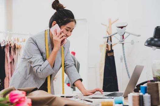 Attractive Asian female fashion designer working in clothing shop studio and using mobile phone to contact customer. Woman receiving order from incoming call. Tailor sewing. People lifestyle concept