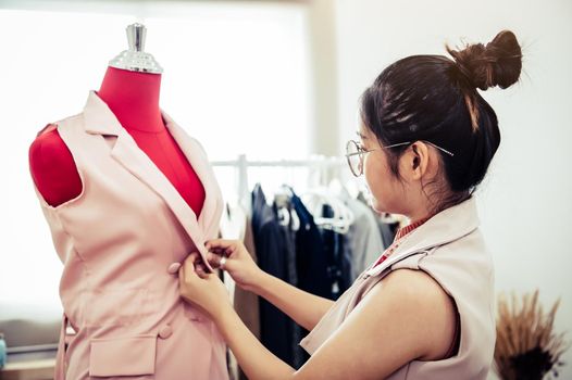 Asian female fashion designer girl making fit on the formal dress uniform clothes on mannequin model. Fashion designer stylish showroom. Sewing and tailor concept. Creative dressmaker stylist.