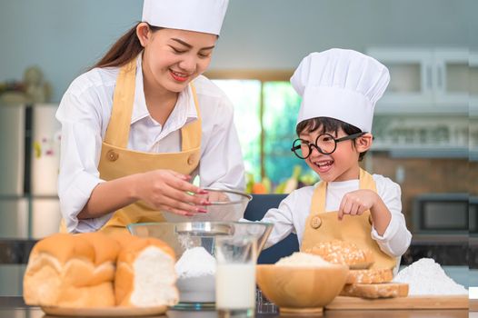 Cute little Asian boy and beautiful mother sifting dough flour with sifter sieve colander in home kitchen on table for prepare to baking bakery and cake. Thai kids playing with flour as chef funny