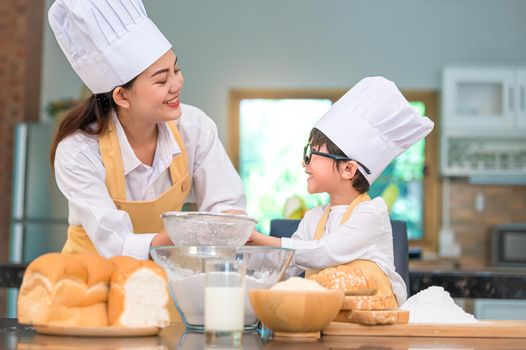 Cute little Asian boy and beautiful mother sifting dough flour with sifter sieve colander in home kitchen on table for prepare to baking bakery and cake. Thai kids playing with flour as chef funny