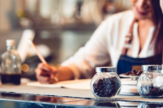 Closeup of coffee bean bottle and happy Female barista write down report note on notebook for calculate business cost and profit as shop owner entrepreneur background in coffee shop. People lifestyle