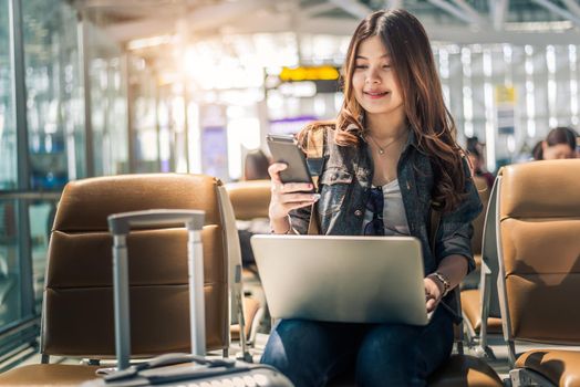 Young Asian female passenger using laptop and smart phone while sitting on seat in terminal hall and waiting for flight in airport . People lifestyles and Happy woman. Technology and Travel concept. 