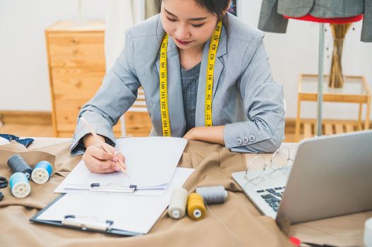 Attractive Asian female fashion designer working in home office workshop. Stylish fashionista woman creating new cloth design collection. Tailor and sewing. People lifestyle and occupation concept