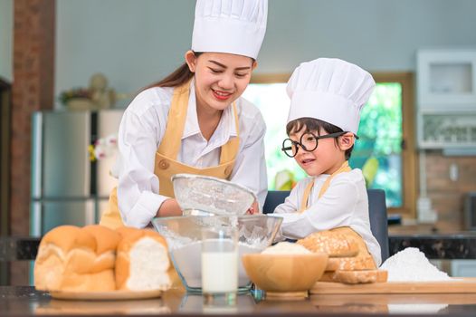 Cute little Asian boy and beautiful mother sifting dough flour with sifter sieve colander in home kitchen on table for prepare to baking bakery and cake. Thai kids playing with flour as chef funny