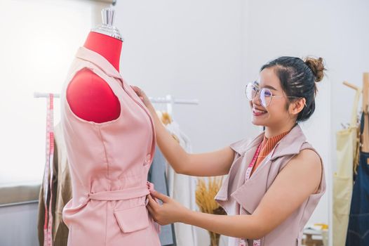 Happy Asian female fashion designer girl making fit on the formal dress uniform clothes on mannequin model. Fashion designer stylish showroom. Sewing and tailor concept. Creative dressmaker stylist.